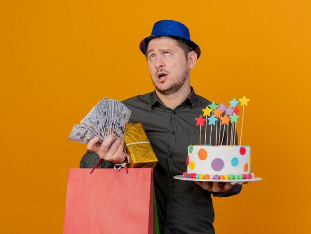 fatigué jeune homme de fête portant un chapeau bleu tenant des cadeaux avec un gâteau et de l'argent isolé sur orange