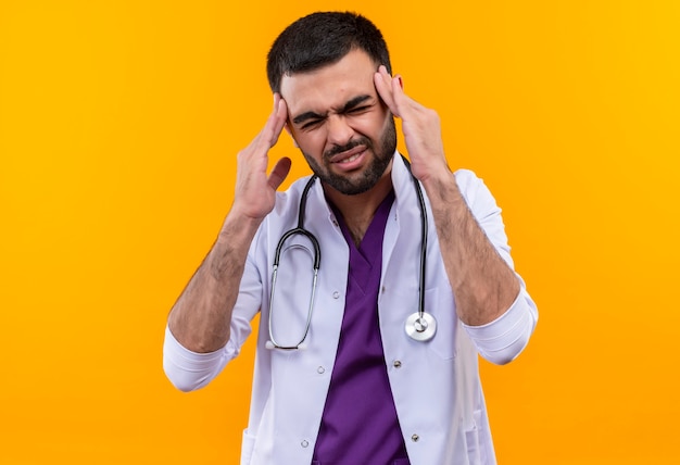 Fatigué de jeune homme médecin portant une blouse médicale stéthoscope a mis ses doigts sur le front sur un mur jaune isolé