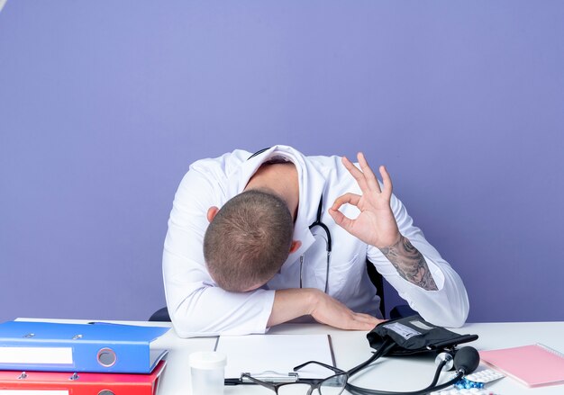 Fatigué de jeune homme médecin portant une robe médicale et un stéthoscope assis au bureau avec des outils de travail mettant la tête sur le bureau et faisant signe ok isolé sur fond violet