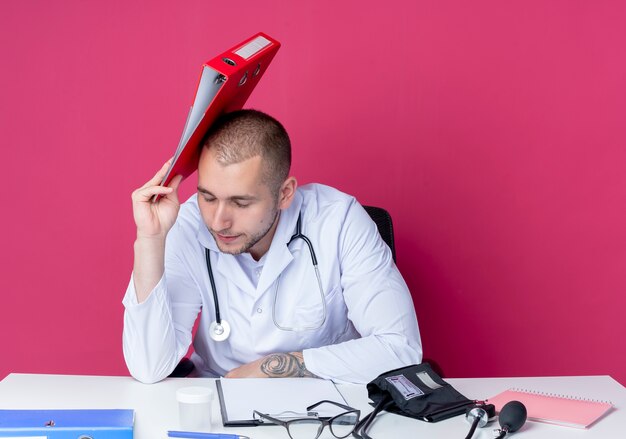 Fatigué de jeune homme médecin portant une robe médicale et un stéthoscope assis au bureau avec des outils de travail tenant le dossier et toucher la tête avec elle avec les yeux fermés isolé sur fond rose