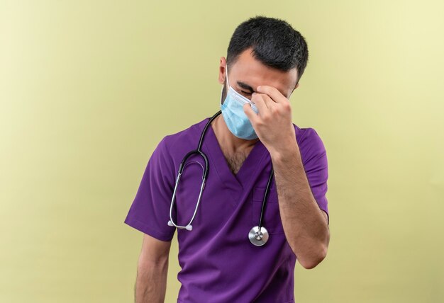 Fatigué de jeune homme médecin portant des vêtements de chirurgien violet et un masque médical stéthoscope a mis sa main sur le front sur un mur vert isolé