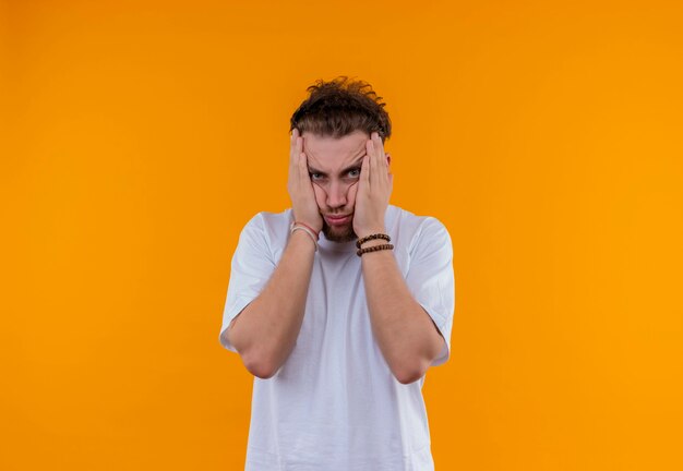 Fatigué de jeune homme portant un t-shirt blanc a mis ses mains sur les joues sur un mur orange isolé