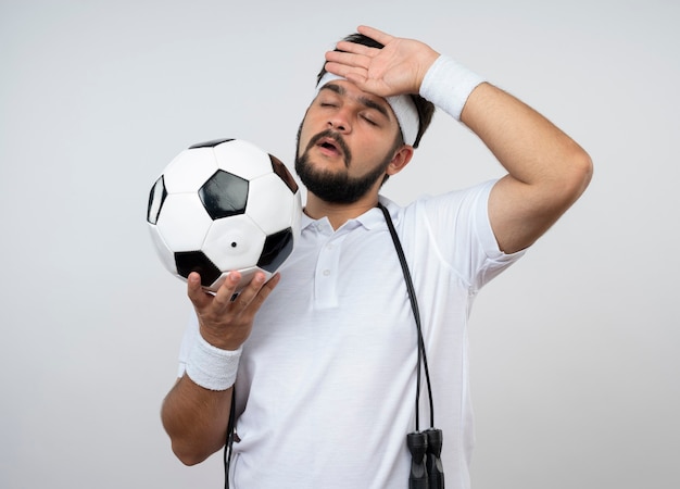 Fatigué de jeune homme sportif aux yeux fermés portant bandeau et bracelet avec corde à sauter sur l'épaule tenant le ballon mettant la main sur le front isolé sur un mur blanc