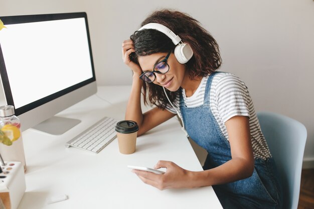 Fatigué de jolie jeune femme assise sur le lieu de travail, tenant la tête et le message texte, tenant le téléphone