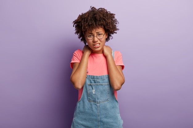 Fatigué mécontent femme sans joie a les cheveux afro garde les deux mains sur le cou, sourit narquoisement de la douleur, être fatigué de travailler à l'ordinateur