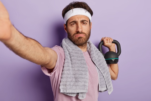 Photo gratuite fatigue sérieux homme fort bodybuilder a des exercices avec du poids, veut avoir des biceps parfaits, démontre la puissance et l'énergie, fait selfie, habillé en tenue de sport, s'entraîne en salle de gym. musculation