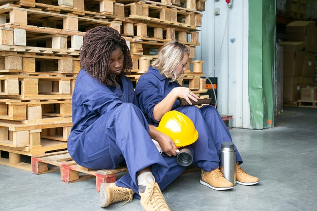 Fatigué somnolent divers ouvriers d'usine assis sur des palettes en bois avec thermos et cookies