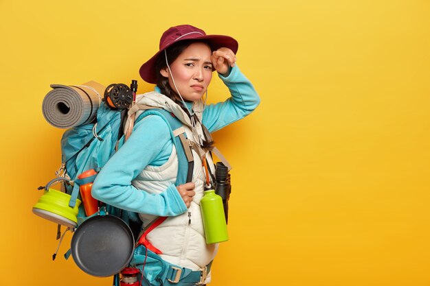 Fatigué touriste épuisé marche longue distance à pied, regarde avec insatisfaction à la caméra, se tient sur le côté contre le mur jaune, porte un sac à dos avec des affaires personnelles