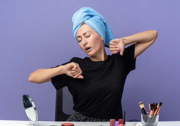 Photo gratuite fatigué avec les yeux fermés jeune belle fille est assise à table avec des outils de maquillage essuyant les cheveux dans une serviette étirant les mains isolées sur fond bleu