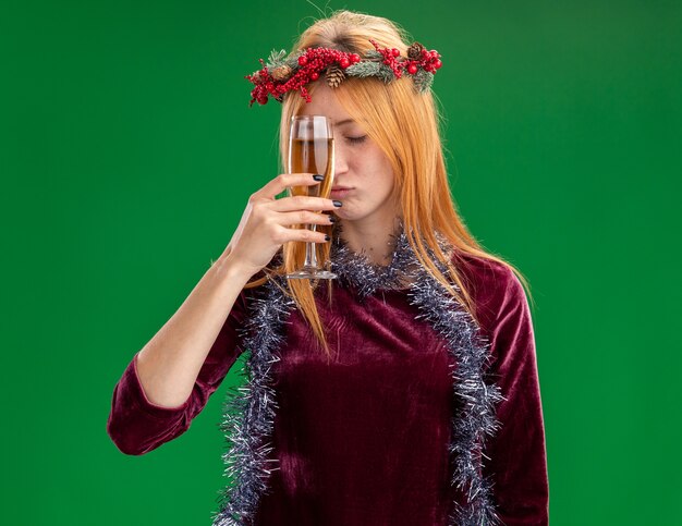 Fatigué avec les yeux fermés jeune belle fille vêtue d'une robe rouge avec une couronne et une guirlande sur le cou tenant un verre de champagne autour du front isolé sur fond vert