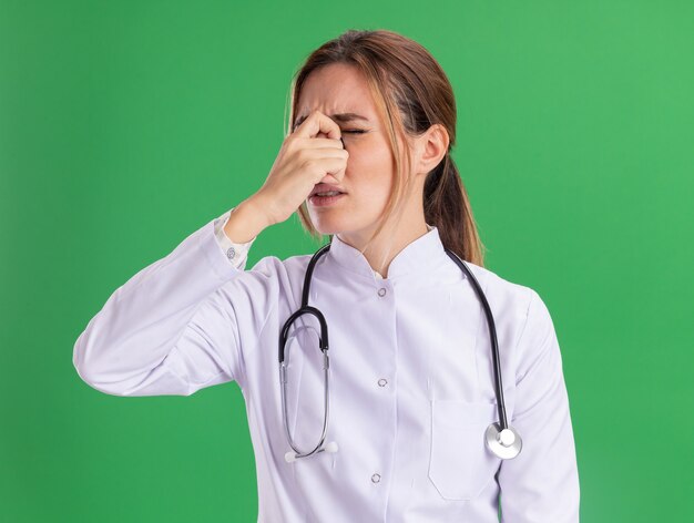 Fatigué avec les yeux fermés jeune femme médecin portant une robe médicale avec stéthoscope a saisi le nez isolé sur mur vert