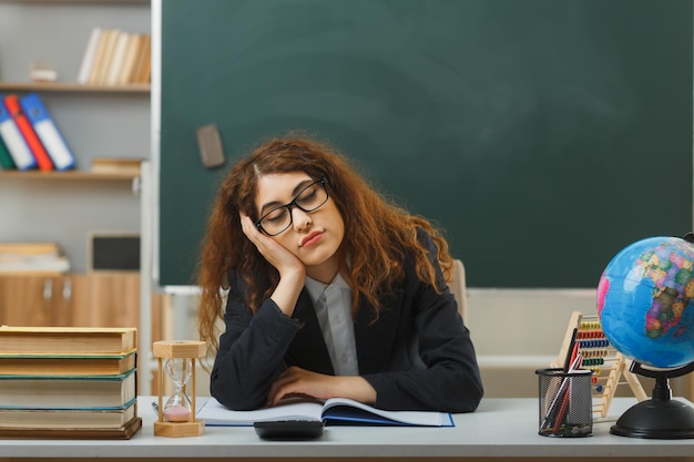 fatigué avec les yeux fermés mettant la main sur la joue jeune enseignante portant des lunettes assis au bureau avec des outils scolaires en classe