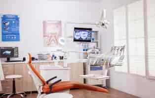 Photo gratuite fauteuil dentaire et autres accessoires utilisés par le dentiste dans une armoire vide. cabinet de stomatologie avec personne dedans et équipement orange pour le traitement oral.