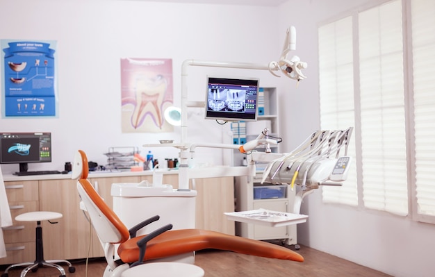 Fauteuil dentaire et autres accessoires utilisés par le dentiste dans une armoire vide. Cabinet de stomatologie avec personne dedans et équipement orange pour le traitement oral.