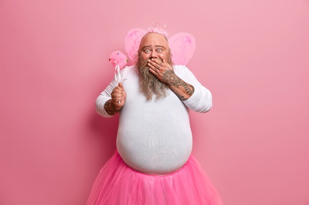 Une fée masculine barbu positive glousse sur son image amusante, a une capacité incroyable à faire de la magie, porte des ailes, un pull blanc et une jupe plissée, démontre un pouvoir magique