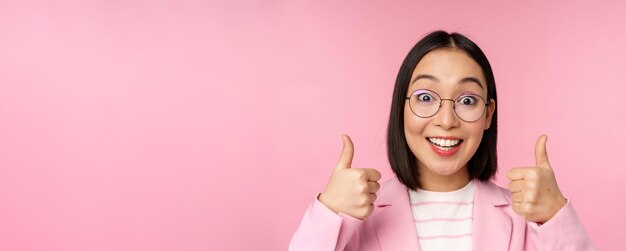 Félicitations géniales Visage d'une femme d'affaires asiatique excitée dans des verres souriant heureux montrant les pouces vers le haut en signe d'approbation debout sur fond rose