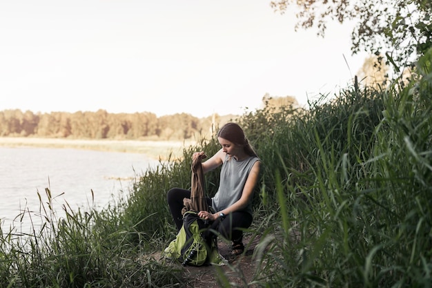 Femme accroupie près du lac retirant les vêtements du sac à dos