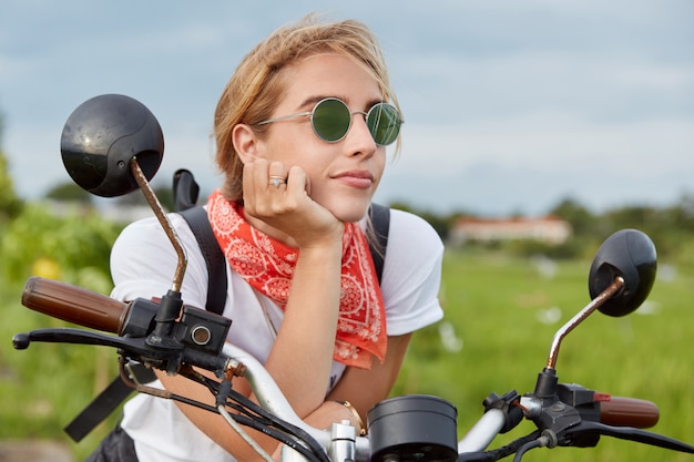 Femme active pensive regarde avec une expression réfléchie à distance alors qu'elle est assise sur une moto, prend une pause après une longue conduite, pose sur les transports en plein air, apprécie la haute vitesse et la nature merveilleuse