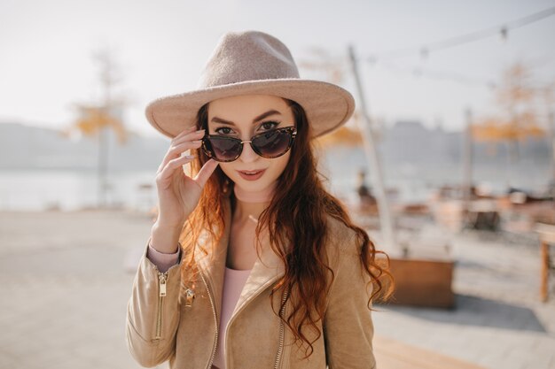 Femme adorable aux cheveux roux ondulés touchant ses lunettes de soleil