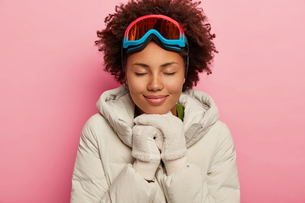 Femme adorable en bonne santé aux cheveux bouclés, porte un manteau matelassé en duvet blanc et des mitaines, utilise un masque de ski, les yeux fermés