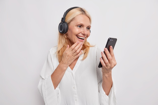 Une femme adulte aux cheveux blonds et heureuse passe un appel en ligne utilise un smartphone et un casque, heureuse d'entendre que son meilleur ami apprend une nouvelle technologie regarde une vidéo sur un appareil mobile isolé sur un mur de studio blanc