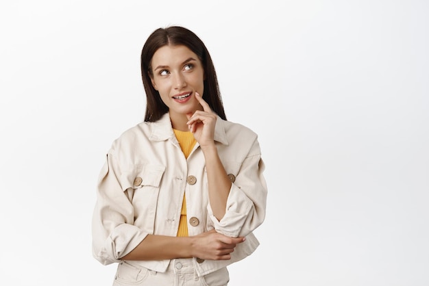 Femme adulte intriguée faisant le choix d'imager qch intéressant en levant les yeux pensif et souriant avoir qch à l'esprit debout sur fond blanc et pensant