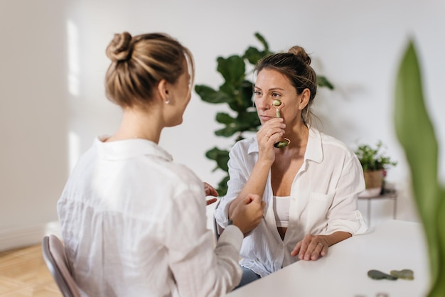 Photo gratuite une femme adulte à la peau claire utilise un masseur à rouleaux tout en étant assise à côté de son esthéticienne dans un studio lumineux. concept de soins du visage spa à domicile