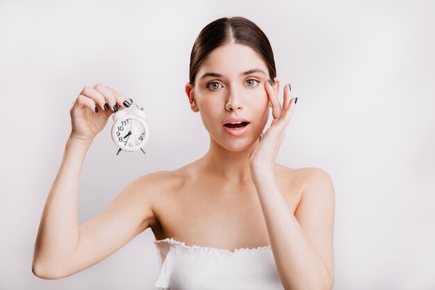 Femme adulte avec une peau saine élastique, tenant un petit réveil blanc.