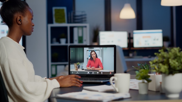 Femme d'affaires africaine discutant avec une femme partenaire à distance en ligne assise devant un ordinateur portable travaillant dans un bureau de démarrage parlant par appel vidéo lors d'une réunion virtuelle à minuit, à l'aide d'un casque