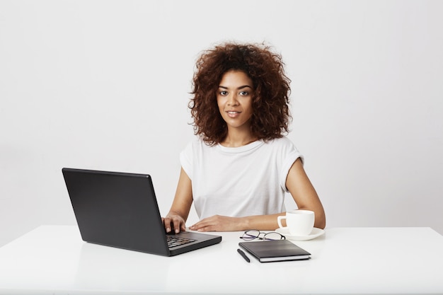 Femme d'affaires africaine souriant assis sur une table au lieu de travail sur un mur blanc.