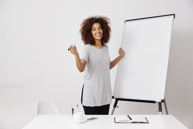 Femme d'affaires africaine souriant debout près d'un tableau blanc sec vierge dans un espace ouvert de bureau, expliquant son idée d'application ou un plan d'affaires sur un mur blanc.