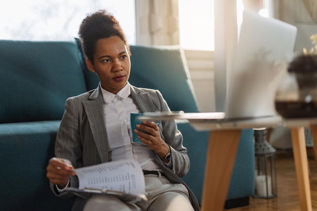 Femme d'affaires afro-américaine examinant des rapports et utilisant un ordinateur portable tout en travaillant à la maison