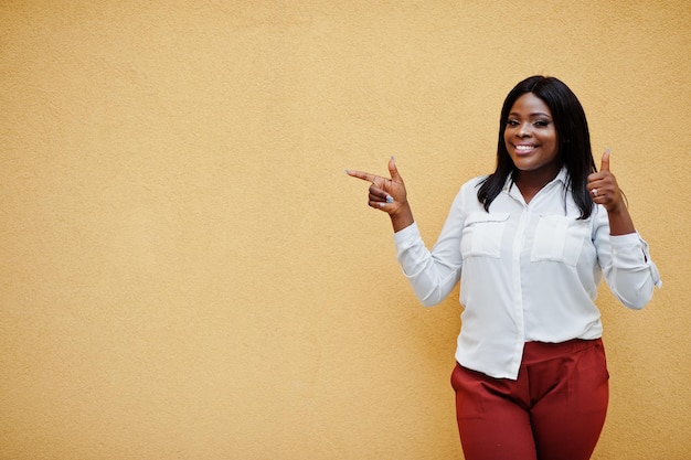 Photo gratuite femme d'affaires afro-américaine formellement habillée en chemisier blanc et pantalon rouge isolé sur fond jaune et montre le doigt vers l'espace libre