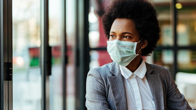 Femme d'affaires afro-américaine portant un masque de protection sur son visage assise dans un café et regardant par la fenêtre