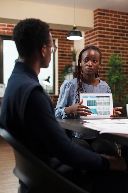 Femme d'affaires afro-américaine présentant à un collègue des données statistiques sur la documentation comptable sur tablette. Le comptable est informé par le chef de projet des revenus et dépenses déclarés à tort.
