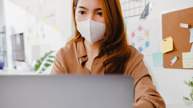 Une femme d'affaires asiatique porte un masque facial pour la distanciation sociale dans une nouvelle situation normale pour la prévention des virus tout en utilisant la présentation d'un ordinateur portable à des collègues sur le plan en appel vidéo pendant le travail au bureau.