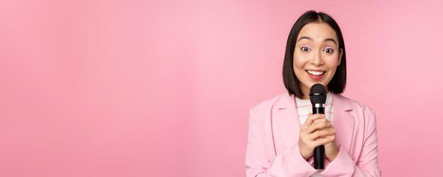 Femme d'affaires asiatique prononçant un discours tenant un microphone et souriant debout en costume sur fond rose