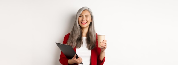 Photo gratuite femme d'affaires asiatique réussie buvant du café et faisant un clin d'œil joyeux à la caméra debout avec clipboar