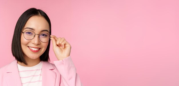 Femme d'affaires asiatique réussie dans des verres souriant et regardant un appareil photo professionnel portant un costume debout sur fond rose