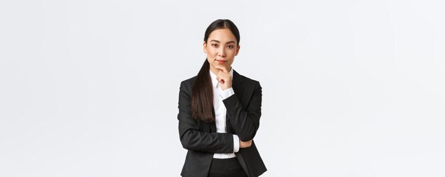 Une femme d'affaires asiatique sérieuse et ravie a une idée intéressante de toucher le menton et de regarder la caméra avec ruse en se tenant debout en costume sur fond blanc
