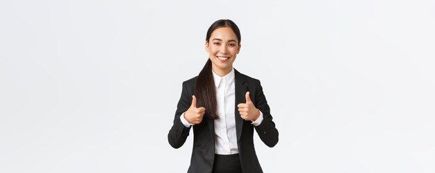 Femme d'affaires asiatique souriante et confiante, professionnelle, assure que tout se passe bien en montrant le pouce levé, recommande une agence ou un produit comme et approuve, dit bravo, bon travail ou bon travail