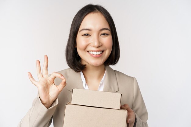 Femme d'affaires asiatique souriante montrant un signe correct et des boîtes avec des marchandises de livraison préparent la commande pour le client debout sur fond blanc