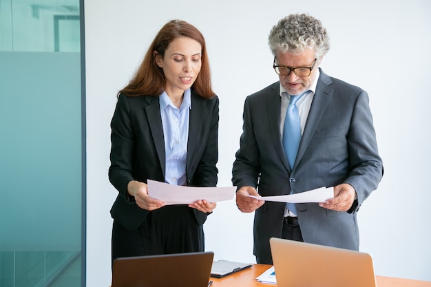 Femme d'affaires aux cheveux roux expliquant le projet, tenant du papier et debout près de la table avec des ordinateurs portables