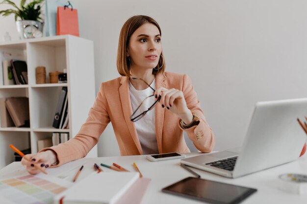 Femme d'affaires aux yeux bruns en veste lumineuse pose assis au bureau dans un bureau blanc