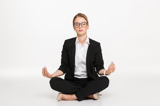 Femme d'affaires blonde calme dans la méditation de lunettes en studio avec les yeux fermés sur le mur blanc