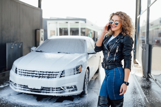 Une femme d'affaires blonde et prospère parle par téléphone près de sa voiture sur une voiture de service de lavage