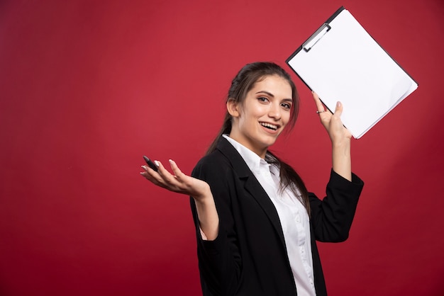 Femme d'affaires brune posant avec presse-papiers sur fond rouge.