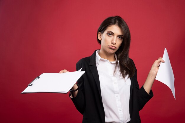 Femme d'affaires brune tenant un tas de documents sur fond rouge.