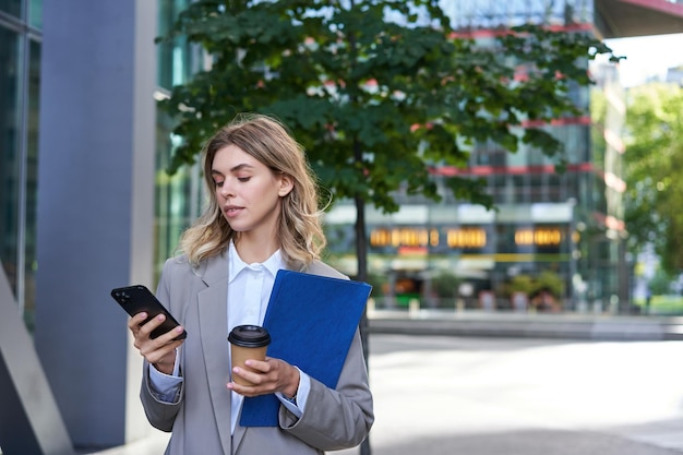 Femme d'affaires buvant du café et tenant des documents avec un smartphone allant travailler au bureau debout