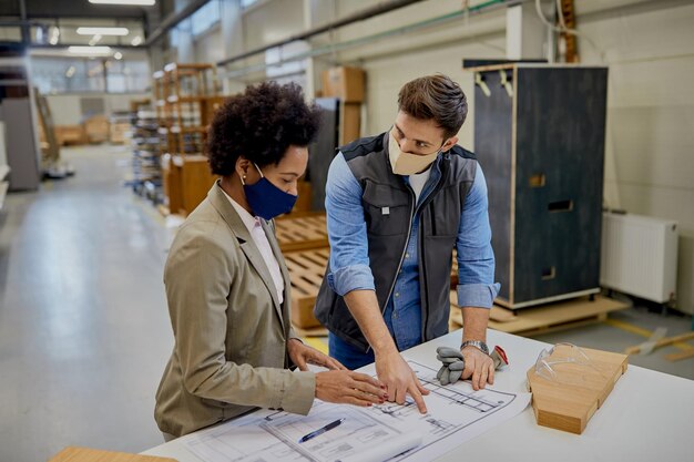 Femme d'affaires et charpentière afro-américaine portant des masques faciaux lors de l'analyse des plans de conception dans l'usine de production